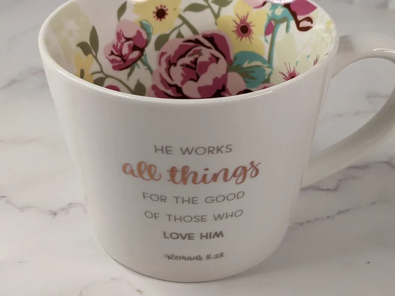 A white ceramic coffee mug with a floral interior design, and a Bible verse in metallic lettering on the outside.