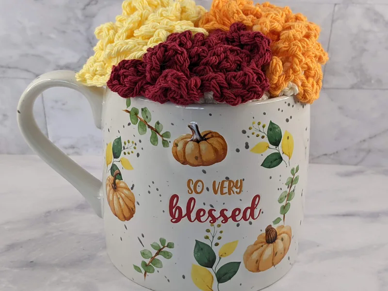 An autumn mug with crocheted dishcloths. The mug says, "so very blessed" and has images of pumpkins.