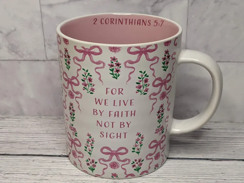 A religious mug that's white with a pink floral design, featuring a pink bow pattern and a Bible verse.