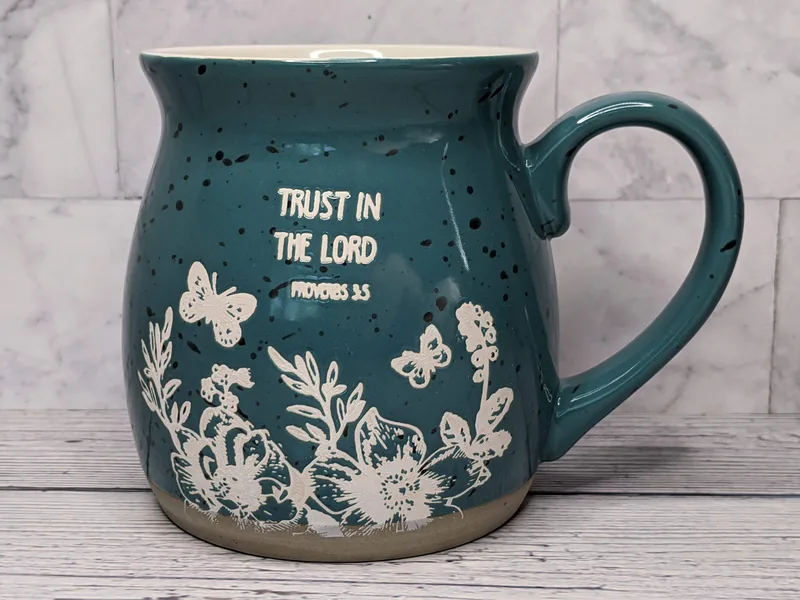 A large teal coffee mug with a Bible verse, Trust in the Lord, and a stamped image of flowers and butterflies.