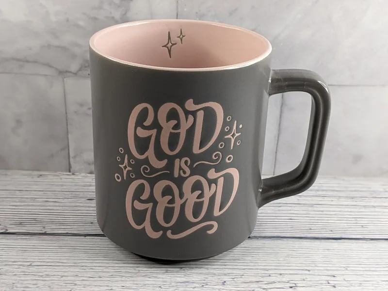 A gray mug with a pink interior, with the words "God is Good" stamped on the exterior in pink lettering.