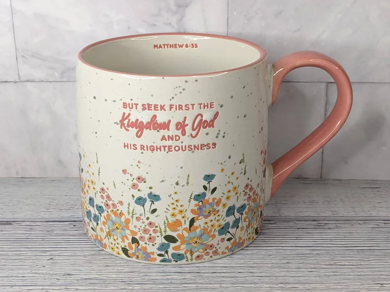 A large white coffee mug with a pink handle, floral design, and a Bible verse inscription about seeking the kingdom of God first.
