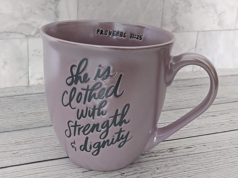A lavender purple mug with the words "she is clothed with strength and dignity" on both sides.