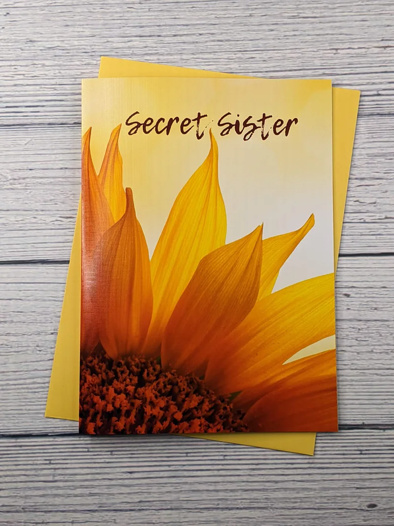 A yellow greeting card with an image of a sunflower and the words Secret Sister, folded on top of a yellow envelope.