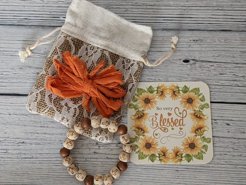 A burlap and lace gift bag with a drawstring closure and a fabric orange bow, with a sunflower gift tag, and a wooden bracelet with a cross design.