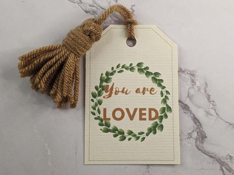 A gift tag that may be used as a bookmark with a yarn tassel and the words: You are loved.