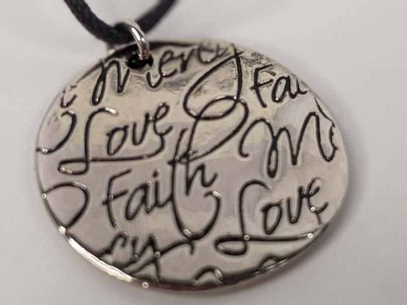 A black-cord necklace with a silver metal pendant featuring the words Love, Faith and Mercy.