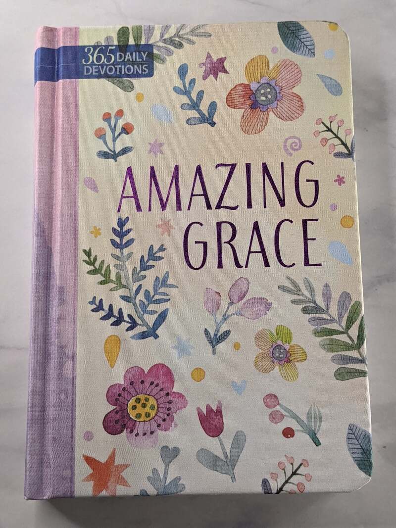 A religious book of devotions with a purple spine, floral design on the cover, and a theme of Amazing Grace.