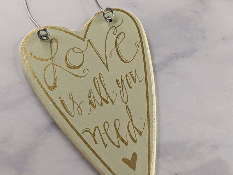 A cream-colored wooden heart cut-out with a wire for hanging it, with the words "love is all you need" in gold lettering.