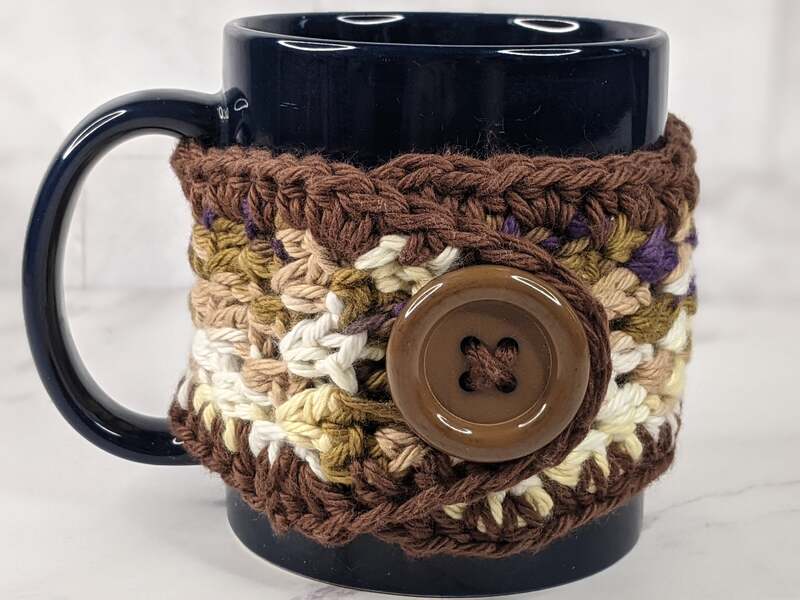 A handmade crocheted brown and tan yarn mug cozy with a plastic brown button closure.