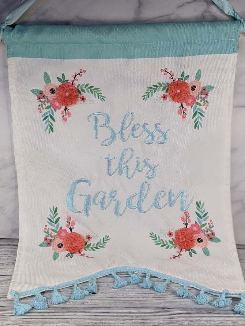 A wall banner that is white fabric with pink flowers embroidered and the words "Bless this garden" in blue.