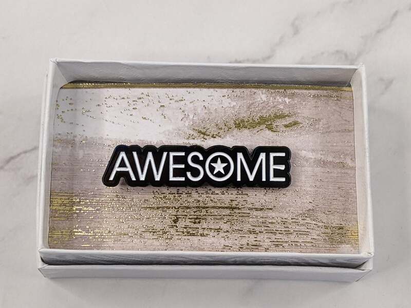 A black lapel pin with white lettering, featuring the word "awesome."