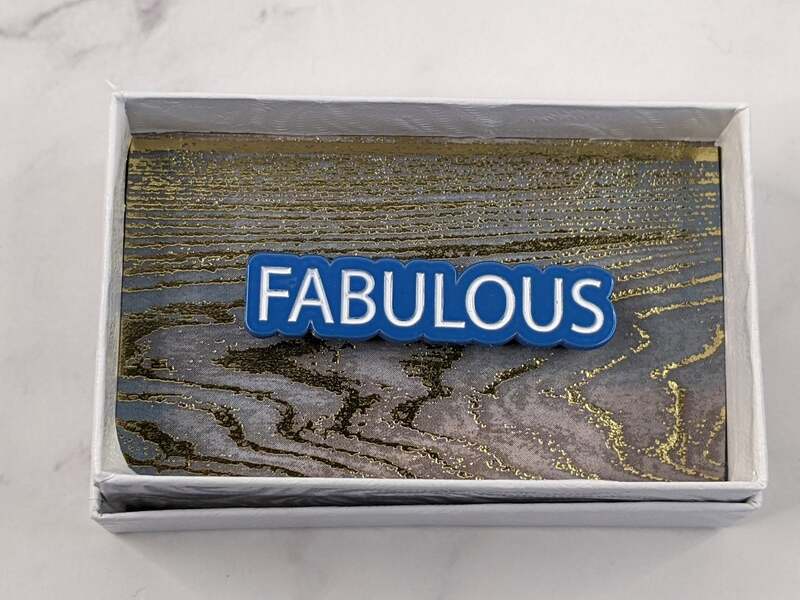 A blue lapel pin with white lettering, featuring the word "fabulous."