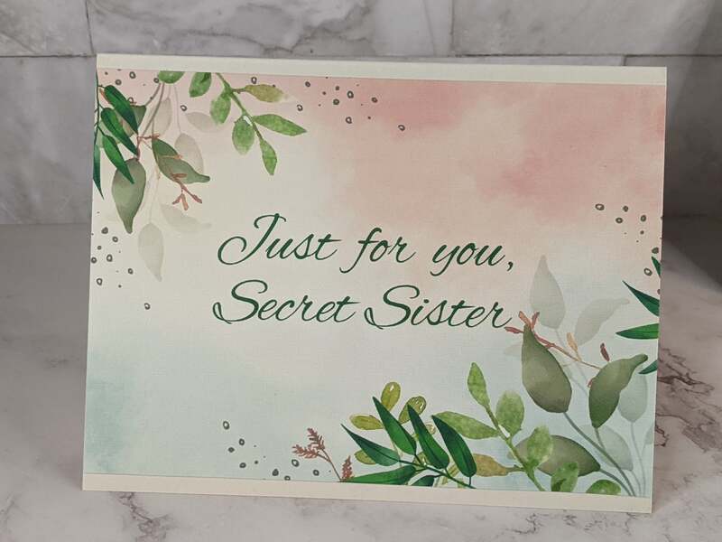 A greeting card for a Secret Sister, wishing her an amazing day, with a pink watercolor design and a green leaf pattern.