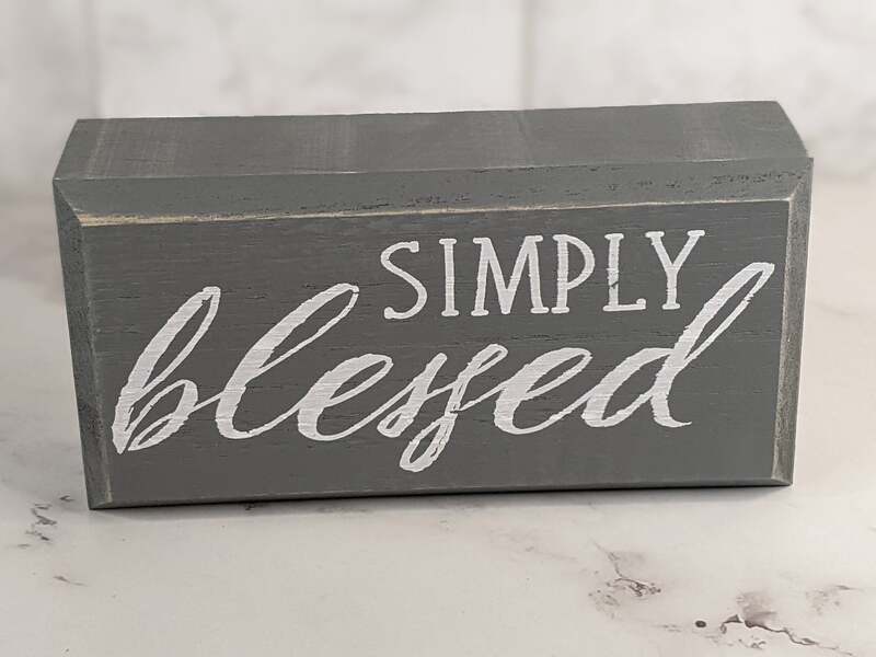 A gray wooden sign that sits on a shelf, with white painted lettering that says, "simply blessed."