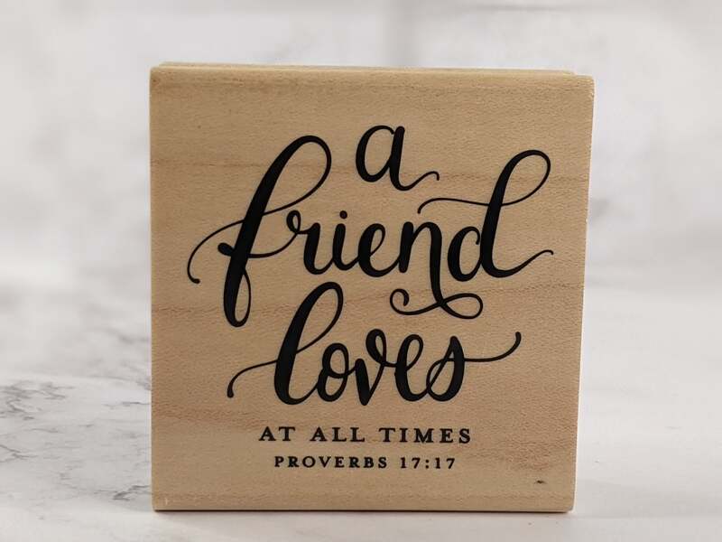 A religious rubber stamp mounted on a wooden block with the Bible verse: A friend loves at all times.